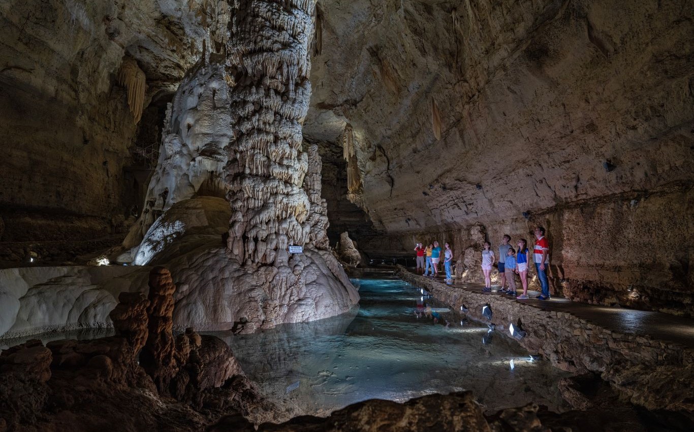A tour group of families in a show cave viewing giant formations in a huge chamber with beautiful water around the columns.